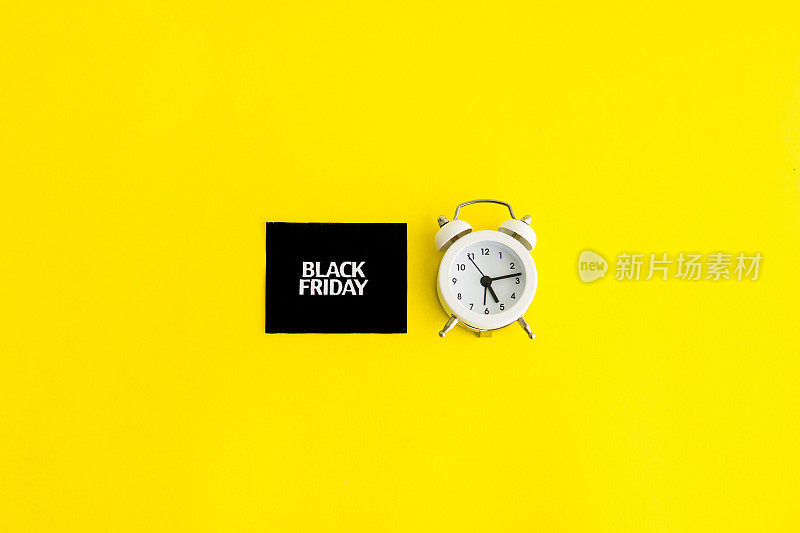 An inscription on a white alarm clock and black square - Black Friday on a yellow background. The topic of sales.